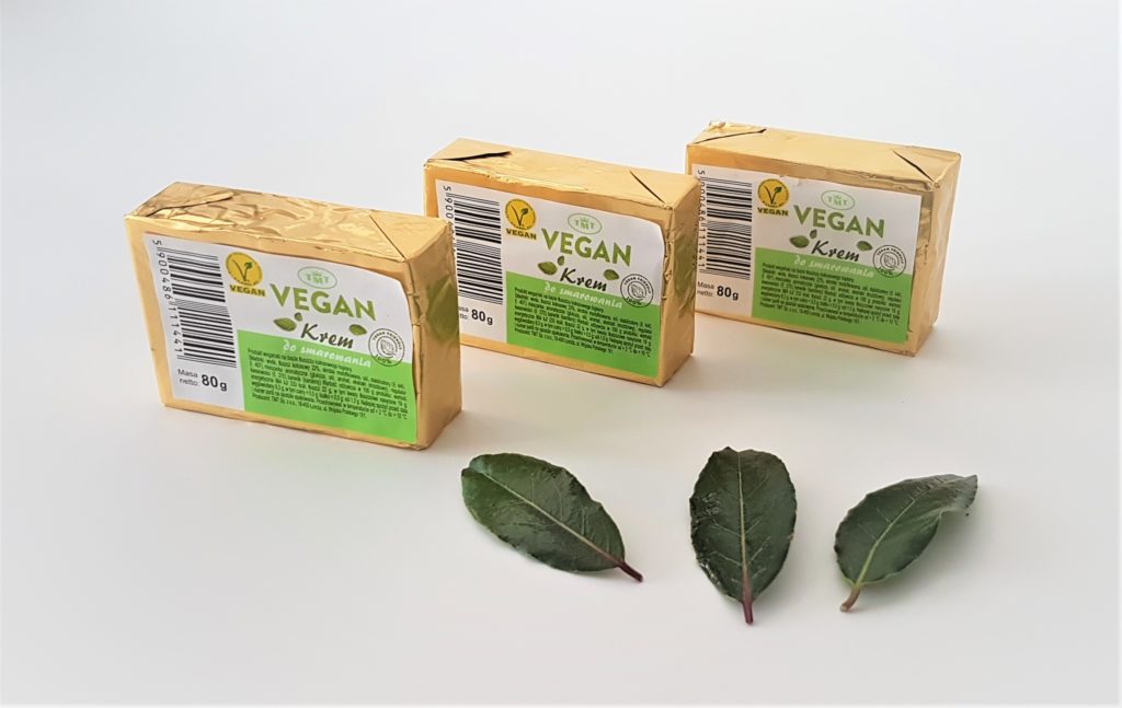 Trzy prostokątne kostki wegańskiego kremu do smarowania marki TMT o masie 80 g każda, zapakowane w złotą folię, ustawione obok siebie na białym tle. Na pierwszym planie leżą trzy ciemnozielone liście laurowe.