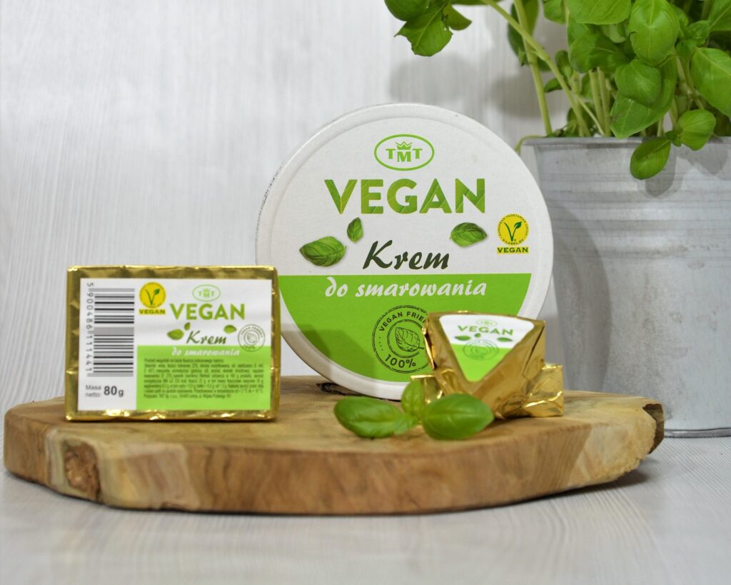 Trzy formy wegańskiego kremu do smarowania marki TMT: kostka 80 g, okrągłe pudełko oraz trójkątna porcja w złotym zawinięciu, ułożone na plastrze drewna. W tle po prawej stronie widoczna bazylia w szarej doniczce, a na pierwszym planie pojedynczy listek bazylii.