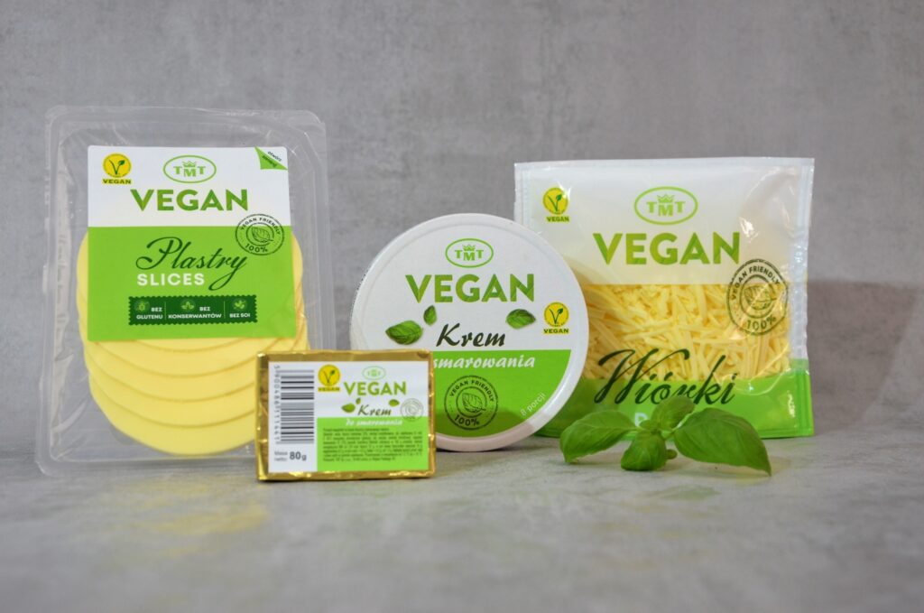 Zestaw czterech produktów wegańskich marki TMT na szarym tle. Od lewej: wegańskie plastry w przezroczystym opakowaniu, kostka kremu do smarowania 80 g w złotym papierku, okrągłe pudełko kremu do smarowania oraz wegańskie wiórki w stojącej torebce. Obok produktów leży świeży listek bazylii.