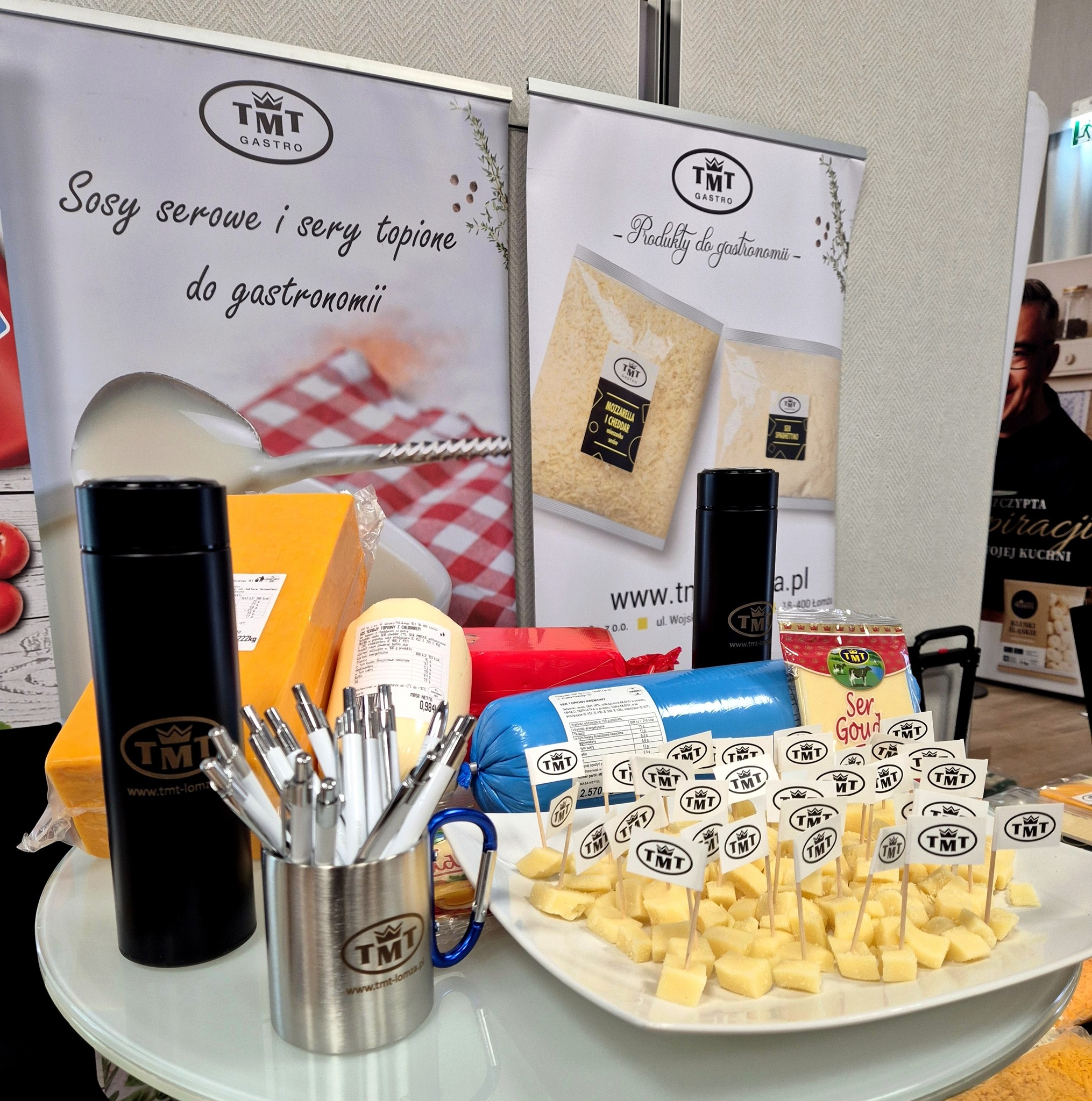 TMT Gastro's promotional stand at the trade fair. In the foreground, a plate of cheese cubes for tasting, with flags featuring the TMT logo, and gadgets: a mug with pens and black thermoses. In the background, blocks of cheese (including one wrapped in blue foil) and two advertising banners with slogans about products for the food service industry.