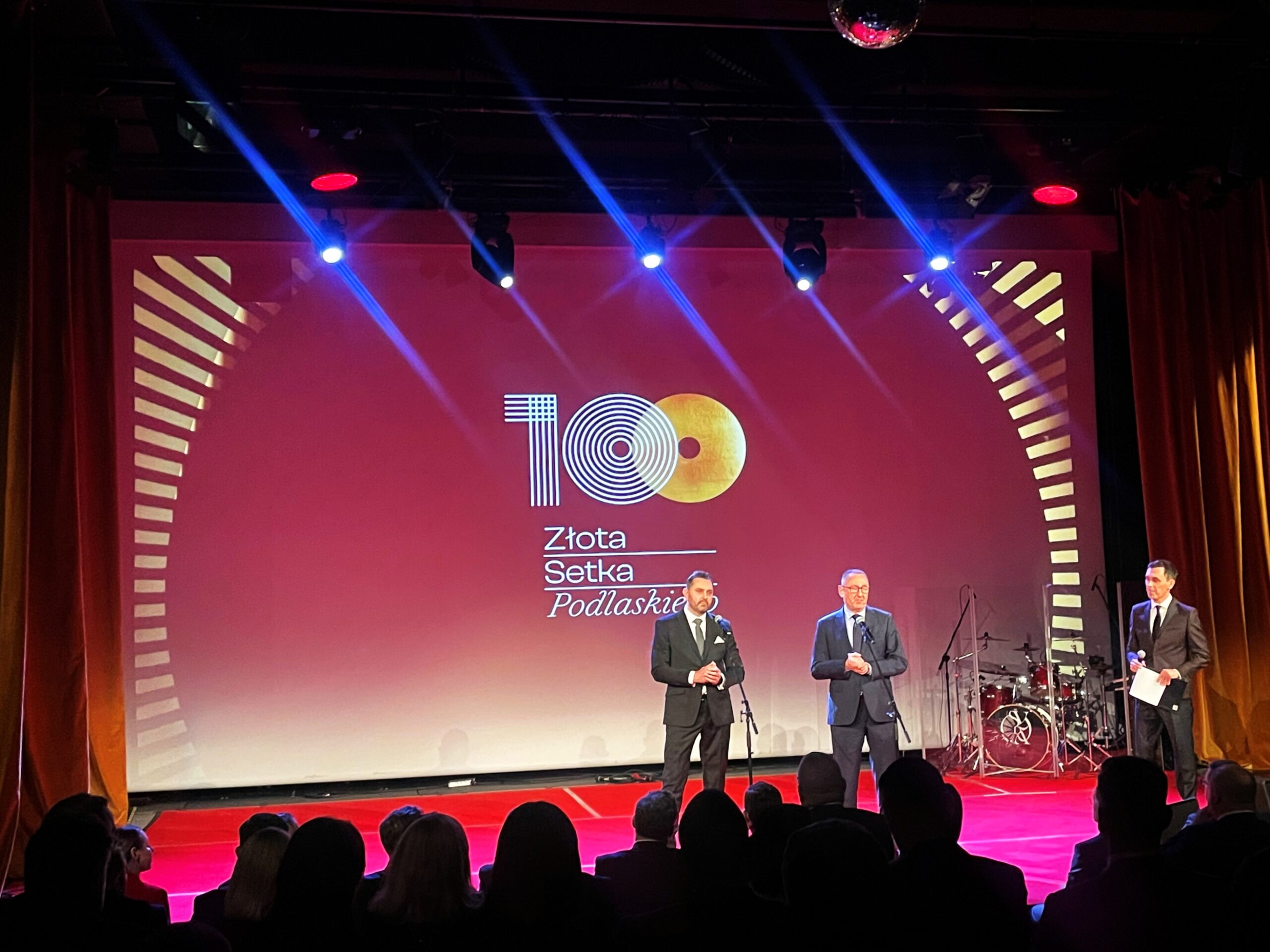 A scene with the "Podlasie Golden Hundred" logo on a red screen. Two men in suits are speaking in front of it.