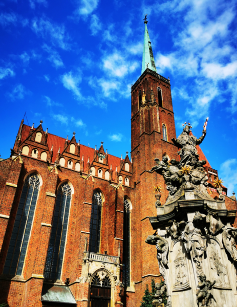 A Gothic red-brick cathedral with a tall tower. Next to it stands a Baroque column with sculptures against a blue sky.