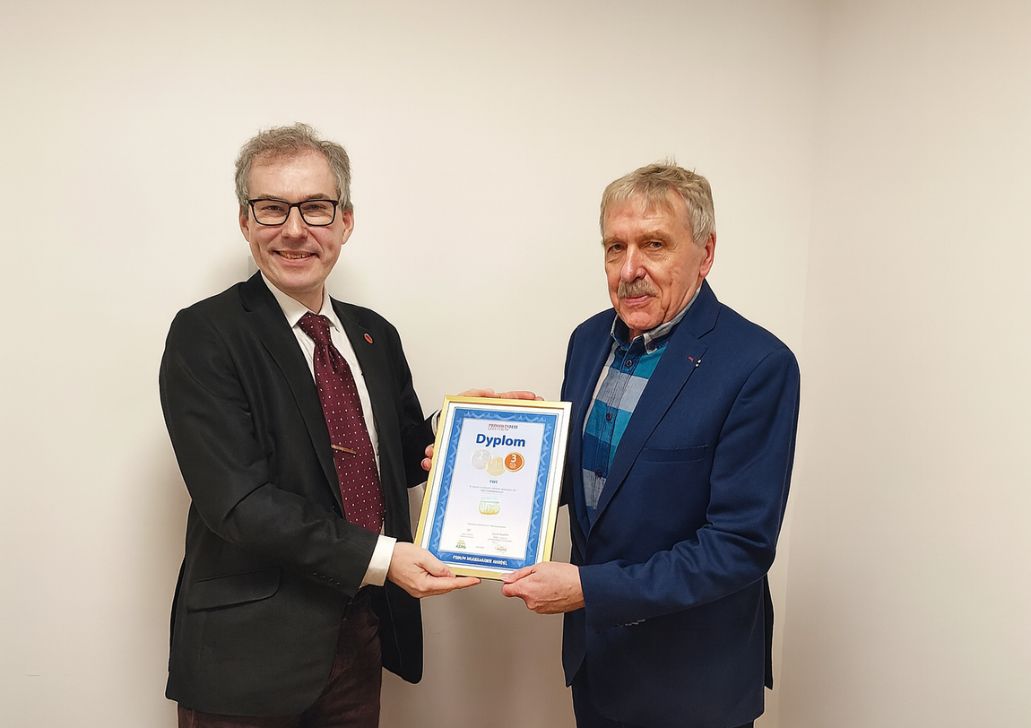 Two middle-aged men stand side by side against a light background, holding a framed document with the large lettering "Diploma" written on it. The man on the left wears glasses, a dark jacket, and a burgundy polka-dot tie, and smiles at the camera. The man on the right wears a navy jacket and a plaid shirt, has a mustache, and stares straight ahead.