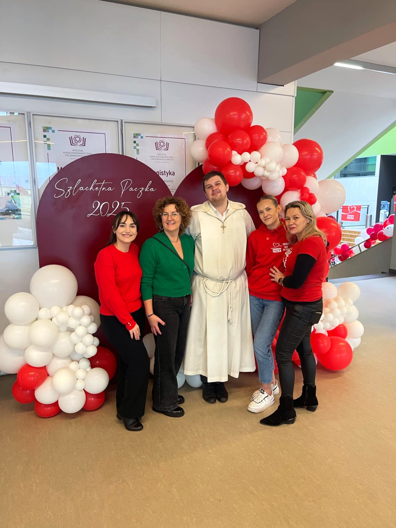 5 osób uśmiechniętych pozuje na bordowej ściancę do zdjęć akcji "Szlachetna paczka 2025". 4 kobiety, pośrodku jeden mężczyzna.