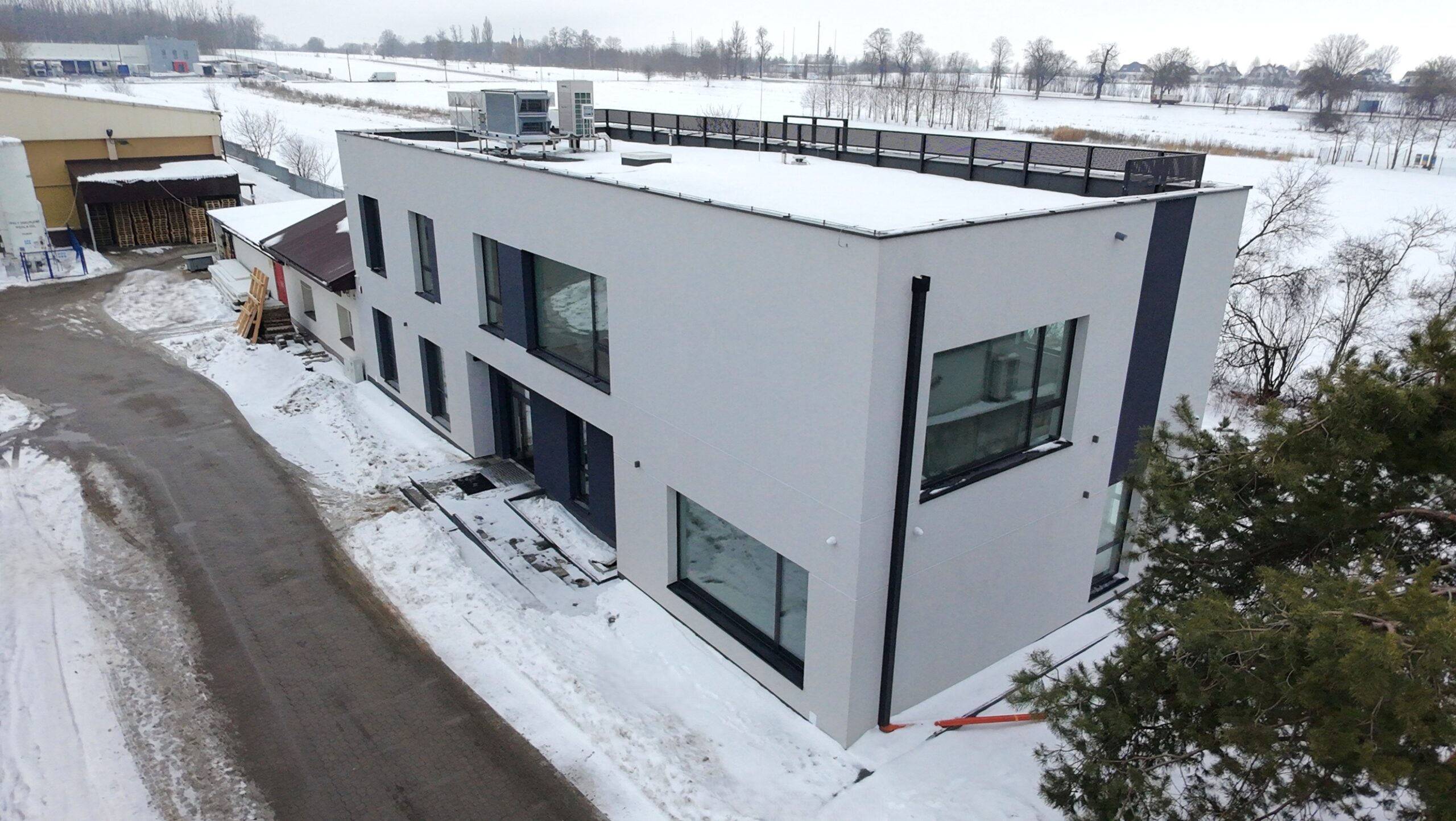 Nowoczesny, dwupiętrowy budynek o białej elewacji z ciemnoszarymi wstawkami i dużymi oknami, otoczony śniegiem. Na płaskim dachu widoczne urządzenia techniczne i barierka. Zdjęcie wykonane z góry, z perspektywy drona.