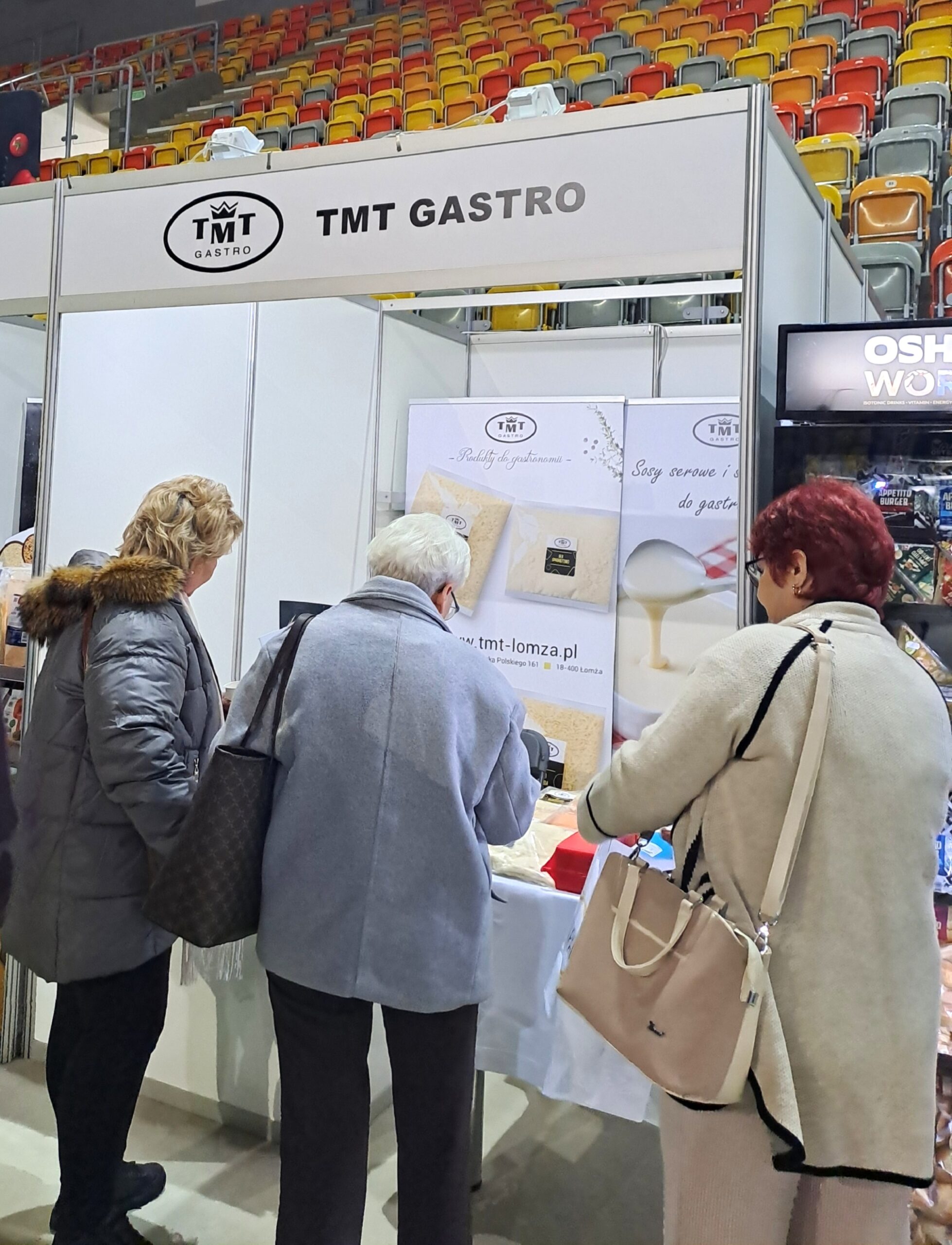 The photo shows a TMT GASTRO exhibition stand during a trade fair held in a sports arena. Three women are standing with their backs to the camera, browsing products on the counter. Above them is a white sign with the company’s black logo and name. In the background, empty, colorful arena stands are visible (yellow, orange, and red seats). The booth features banners advertising cheese sauces and catering products, as well as the website address tmt-lomza.pl.