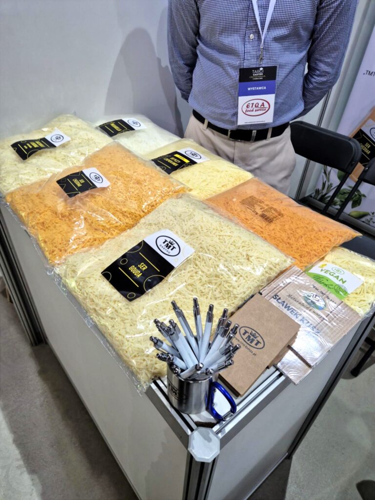 Zbliżenie na biały blat stoiska, na którym wyeksponowano duże, przezroczyste worki z tartym serem w różnych kolorach (m.in. żółta Gouda, pomarańczowy Cheddar oraz białe wiórki wegańskie). Obok leżą gadżety firmowe: metalowy kubek wypełniony biało-niebieskimi długopisami oraz stos brązowych notesów z logo TMT. W tle widać fragment sylwetki wystawcy w niebieskiej koszuli z identyfikatorem "Targi Gastro Częstochowa".