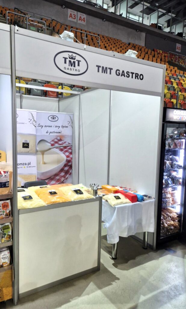 Szerokie ujęcie stoiska TMT GASTRO w hali sportowej na tle pustych, pomarańczowo-żółtych trybun. Stoisko składa się z białych ścianek systemowych i dwóch blatów, na których ułożono produkty serowe. Na lewej ścianie wisi wysoki baner z apetycznym zdjęciem lejącego się sosu serowego i napisem "Sosy serowe i sery topione do gastronomii". Po prawej stronie stoiska stoi wysoka lodówka przeszklona z napojami i przekąskami.