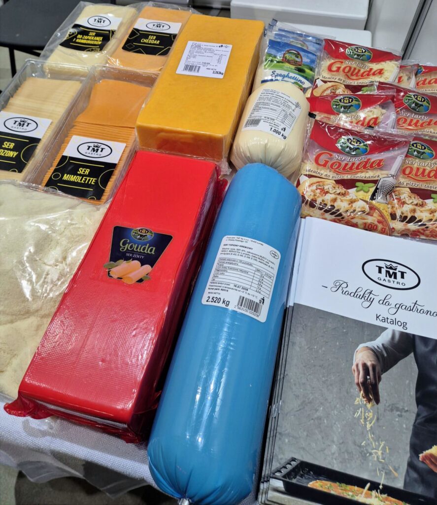 Zdjęcie przedstawiające bogaty wybór produktów serowych TMT GASTRO. Na blacie widoczne są sery w plastrach (Mimolette, Cheddar, do hamburgerów), sery w dużych blokach (czerwone i żółte opakowania), ser topiony w formie niebieskiego „batonu” o wadze 2,5 kg oraz małe opakowania tartej goudy. W prawym dolnym rogu leży katalog firmowy z napisem „Produkty do gastronomii”.