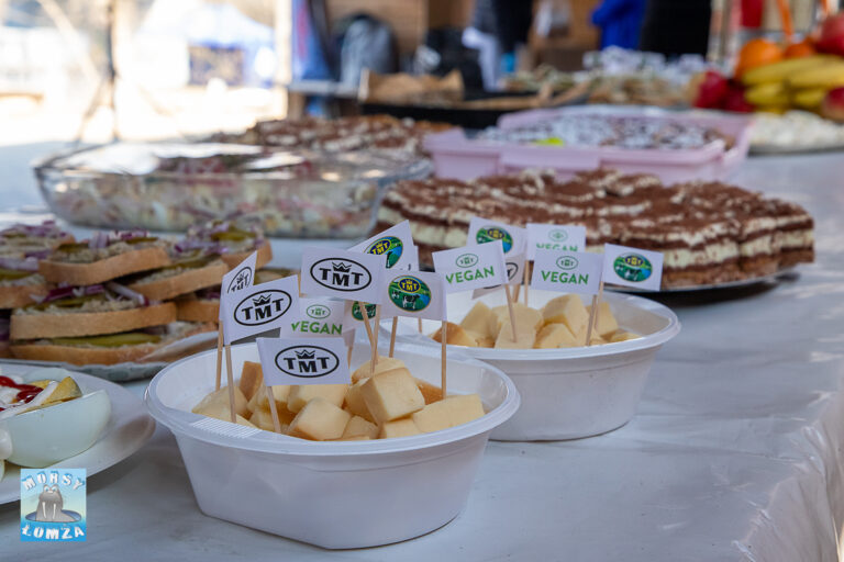 An outdoor snack buffet. In the foreground, white bowls contain cubed cheese marked with small flags featuring the TMT logo and the word VEGAN. Sandwiches, cakes, and fruit are visible in the background.
