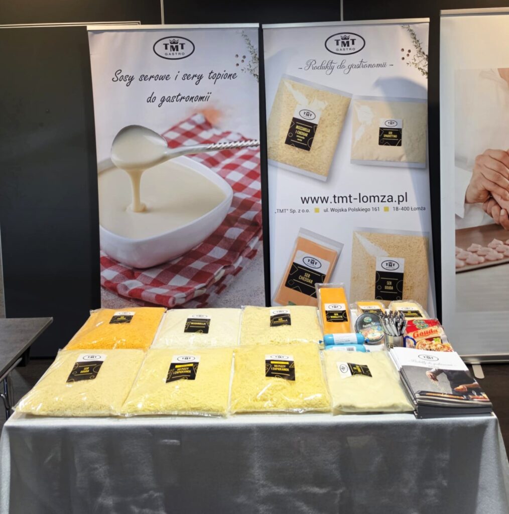 A photo of the TMT Gastro trade show booth, designed in collaboration. In the foreground is a white table displaying products: large bags of shredded cheese (including Gouda, Mozzarella, and Mixarella), block cheeses (Cheddar, Mimolette), and company catalogs. In the background are TMT Gastro advertising roll-ups showcasing their selection of cheese sauces and cheeses for the foodservice industry.