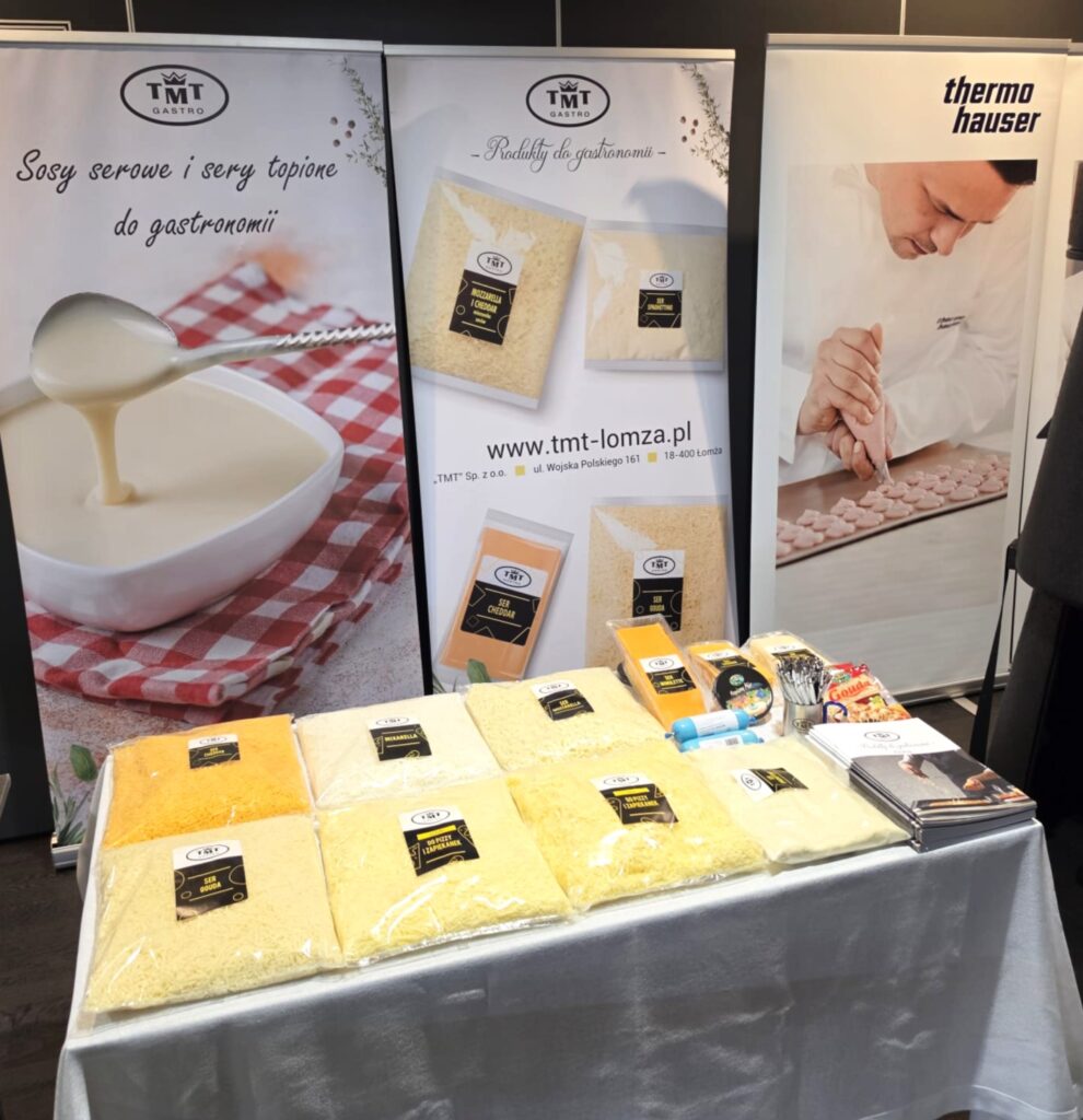 A photo of the TMT Gastro trade show booth, prepared in collaboration. In the foreground is a white table displaying products: large bags of shredded cheese (including Gouda, Mozzarella, and Mixarella), block cheeses (Cheddar, Mimolette), and company catalogs. In the background are TMT Gastro advertising roll-ups featuring their selection of cheese sauces and cheeses for the food service industry.