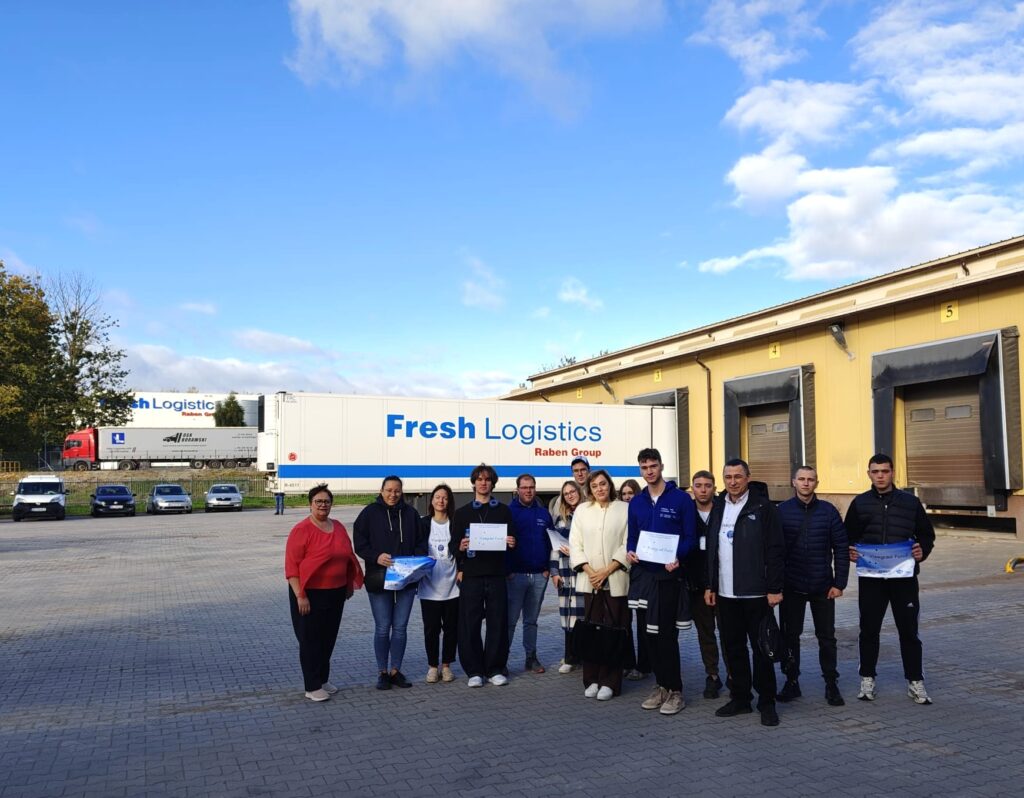Zdjęcie grupowe studentów Wyższej Szkoły Agrobiznesu w Łomży ze Słowacji i Ukrainy podczas wizyty w TMT. Uczestnicy stoją na placu logistycznym przed halą magazynową, na tle samochodów ciężarowych i ramp załadunkowych.